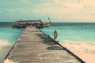A capture of a long and large wooden boardwalk,on a white sand on a Maldivian island, leading to the sea where there's a beautiful gondola moored at the end of it, surrounded by the turquoise seawater