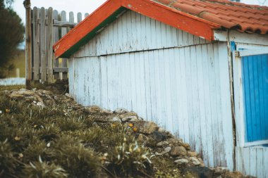 A capture of humble house built of wood painted white, and blue, but with orange tiles in an A shape situated on a small stone slope and undergrowth, the whole property is fenced in with wooden stakes