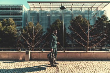 A sophisticated Angolan male was captured outside of the office building, in the back, standing on the pavement wearing a chequered dark green suit and carrying a weekender on a sunny day