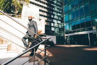 A black white-collar employee moving up the staircase, a metallic railing next to him, the office building with blue windows behind him and he is carrying a weekender bag, wearing dark grey suit