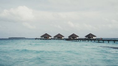 A wide-angle shot of a villa of overwater bungalows and the wooden bridge connecting to the areal in the background, in the foreground the warm turquoise crystal clear sea water