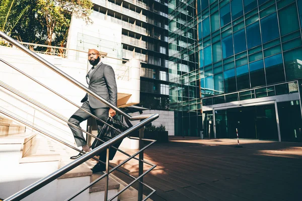 A black white-collar employee moving up the staircase, a metallic railing next to him, the office building with blue windows behind him and he is carrying a weekender bag, wearing dark grey suit