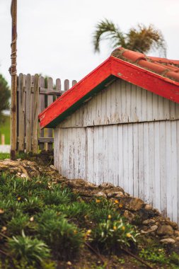 Vertical view of modest wooden house painted in white with orange-red tiles in an A shape situated on a small stone hillside with undergrowth, fenced in with wooden stakes