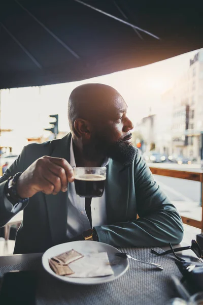 A vertical shot of a handsome black man drinking coffee in a street cafe on a sunny morning; the portrait of a dapper African American guy looking aside while having a coffee in an outdoor restaurant