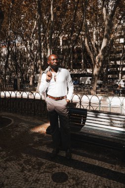 A vertical shot of an elegant mature bald African American man in a white shirt and eyeglasses, with a well-groomed black beard, standing next to a park bench, holding a jacket slung over his shoulder