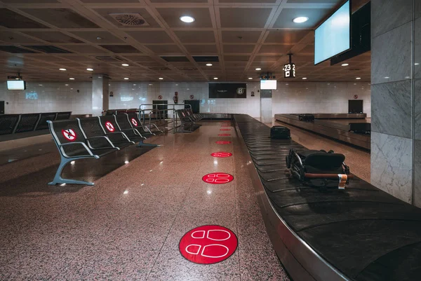 An empty baggage claim zone of a modern airport terminal with rows of seats and different luggage on the conveyor belt; red markers on the marble floor and chairs for a social distancing