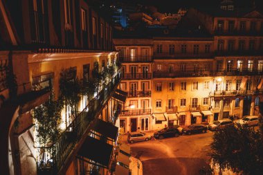 A low key long exposure capture of the view from a residential building balcony, in the middle of the night; The facade of the neighboring apartments with fenced balconies with trees and climbers