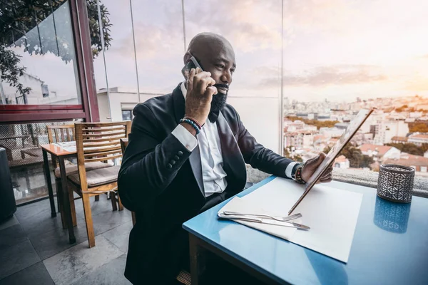 A stylish bald unshaven black male dressed in a sleek black suit is engaged in a phone conversation while perusing the menu at a restaurant located on a terrace overlooking a bustling cityscape