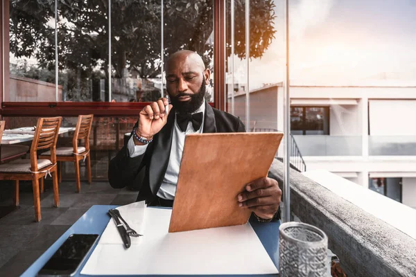 A bald black man dressed in a sleek black tuxedo and bow tie is perusing the cork color menu at a restaurant, located on a rooftop, with a side view while holding a pen; he is looking thoughtful