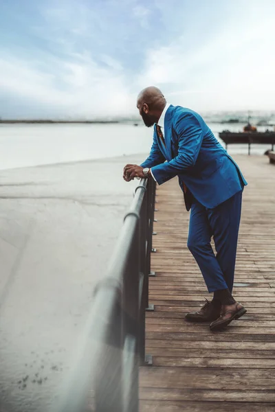 Vertical view of a well-dressed bald black man in a navy blue suit leaning on the iron railing of a wooden river walkway looking in the opposite direction of the camera on a day with some white clouds