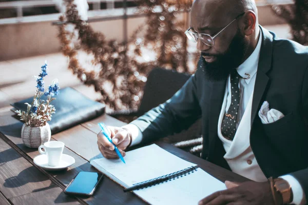 An African writer with a full-grown beard sitting on an outdoor esplanade, dressed in a dark suit, polka-dot tie, and a white vest, reviews his notes, is holding a pen, and while taking a coffee