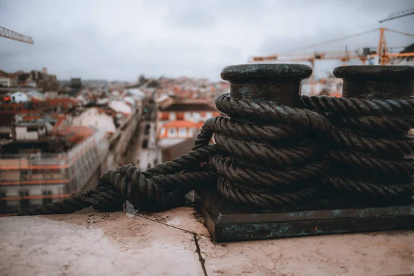 A close-up shot with a focus on a metal sculpture, in medium bronze color, of a bollard with a thick rope attached and around it, with the city of Lisbon out of focus in the background