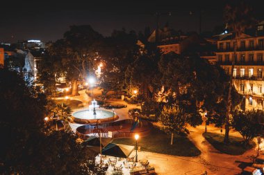 A top view in low-key and long exposure to a lit urban garden at night. In the center, a fountain, a little playground with a slide, a terrace with tables and sunshades, and trees surround the site