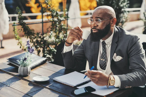 A black balding corporate man with a beard sitting in an outdoor cafe, dressed in a fancy suit, polka-dot tie, and a white vest, reviews his notes with a pen, and while taking a sip of his coffee