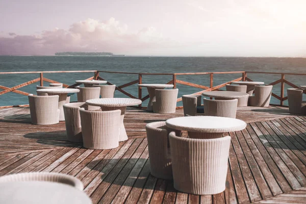 View of an serene outside cafe restaurant area filled by the beach ocean with rattan-effect tables and chairs on the wooden deck and fenced, in a peaceful background with lavender sky with clouds