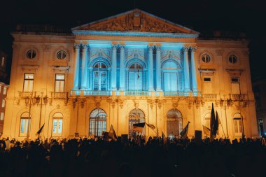 Lisbon, Portugal - February 24, 2023: A night shot of the one-year anniversary of the war protest on a square, as the building behind the protestors is illuminated in the colors of the Ukrainian flag clipart
