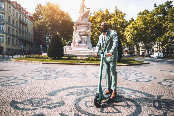 Bald, bearded black man, having a great time while riding an electric scooter whose greenish color matches the suit he is wearing; black man wandering through the city next to a statue and greenery