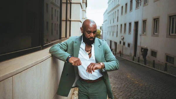 A sophisticated bald black man wearing a pastel green suit and a white shirt, accessorized with necklaces and checks the time on his wristwatch while standing in the middle of a cobblestone street