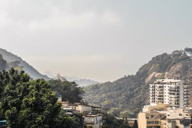 Rio de Janeiro 'da yeşil ağaçlarla kaplı verimli dağlarla çevrili bir daireden manzara, manzarayı çevreleyen mütevazı binalar; güneşli bir güne hafif sis bulutları eşlik ediyor