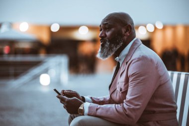 Pembe soluk bir ceket giymiş, kel ve sakallı bir siyah adamın görüntüsü otururken ve telefonunu tutarken dalgın görünüyor. Seçici odaklanma ve geniş bir kopya alanı.