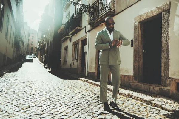 In an old Lisbon neighborhood, a black bald man with a full-grown beard stands confidently in the middle of the street on a sunny day. Wearing a stylish green suit checks the time on his wrist clock