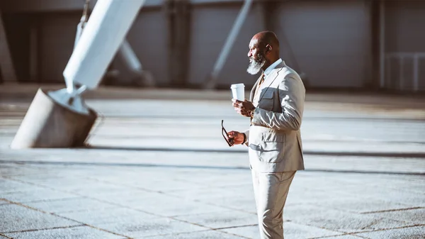 stock image A dapper black businessman in a beige suit is captured in profile against a stunning tensile structure. He holds a cup of coffee in one hand and a pair of glasses in the other, very urban elegantly