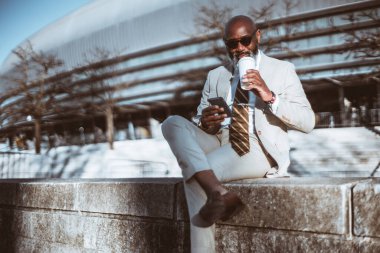 Kel siyah bir adam taş basamaklarda oturuyor, takım elbise giyiyor ve yüzünde içerik ifadesi var. Kahvesini yudumlarken ve telefonundan e-posta okurken bacak bacak üstüne atıyor.