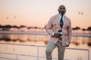 Pembe gökyüzü, Tagus Nehri arkaplanı. Seçici bir şekilde pembe ceketli, beyaz gömlekli, krem pantolonlu ve şık kravatlı kel bir adama odaklan. Elinde kahve ve cep telefonu var. Kopyalama alanı