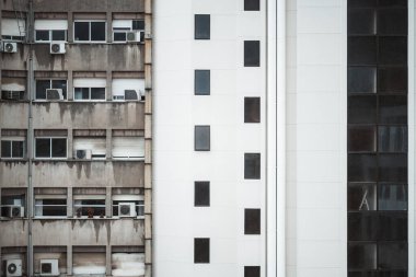 Karşıt cephelerde, solda, birden fazla kliması olan yıpranmış bir daire, sağda ise parlak dikdörtgen pencereli modern bir beyaz konut çarpıcı bir görsel ikilem oluşturur.