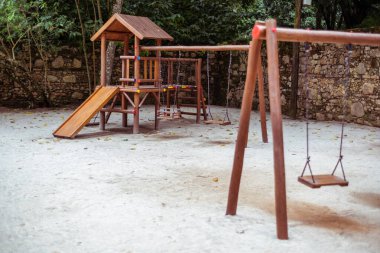 Üçgen çatılı ve çocuk kaydırağıyla küçük ahşap bir eve odaklı tropik bir ortamda bir oyun alanı; çocuklar için kumdaki çeşitli salıncaklar, odakları kesilmiş ön plandakiler.