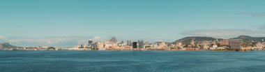 Rio de Janeiro, Brezilya 'nın panoramik manzarası, açık mavi gökyüzüne karşı şehrin ufuk çizgisini gösteriyor. Görüntüde ikonik binalar, yemyeşil tepeler ve Guanabara Körfezi 'nin engin genişliği yer alıyor.