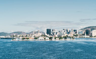 Rio de Janeiro 'nun şehir manzarasının panoramik görüntüsü: modern gökdelenler, tarihi binalar, ve berrak mavi gökyüzü altında yemyeşil tepeler, ön planda Guanabara Körfezi' nin sakin suları hakim
