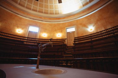 Tarihi bir konferans salonunun içinde yarım daire şeklinde katmanlı ahşap koltuklar var. Odak noktası süslü bir pirinç musluk ve yumuşak ışıklandırmalı kubbeli bir tavanın altında batma