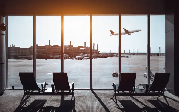 Büyük cam pencerelerden pisti gören bir havaalanı terminali. Bekleme salonunun ön planında dört boş sandalye var ve bir uçağın uzaklıktan havalandığı görülüyor.