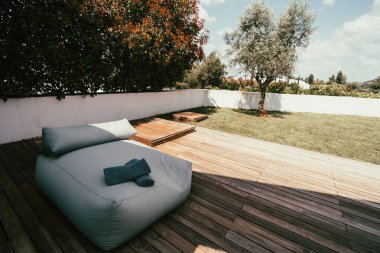 Geniş açılı bir açıyla geniş gri yastıklı rahat bir oturma alanı ve ahşap bir güvertede rulo havlular. Bölge, açık bir gökyüzünün altında bir ağaç ve çalılar da dahil olmak üzere yeşilliklerle çevrili.