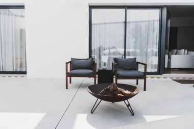 Modern bir bahçe terası: iki gri yastıklı ahşap sandalye ve kütüklü yuvarlak ateş çukuru. Büyük cam sürgülü kapıları ve katıksız perdeleri olan minimalist beyaz bir binaya karşı kurulmuş.