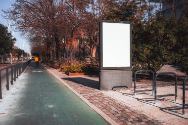 Şehir parkında ağaçlık bir yolda bisiklet yolunun yanında boş bir ilan panosu duruyor. Turuncu ceket giyen bir bisikletçinin silueti uzaklaşıyor ve ön planda bisiklet rafları görülüyor.