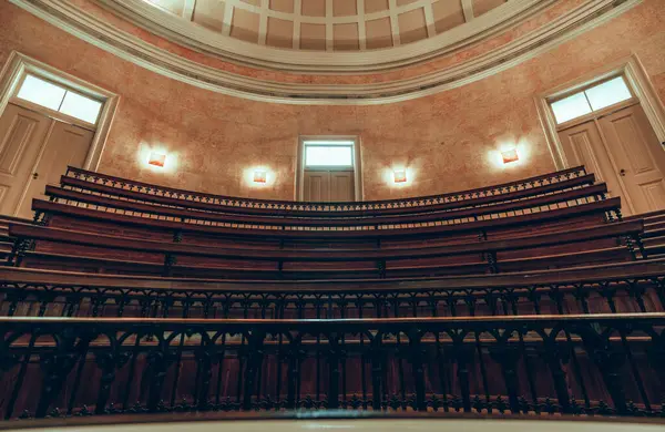 Kavisli ahşap koltukları ve demir parmaklıkları olan eski bir konferans salonunun iç çekimi. Odada sıcak ışıklandırma, dekoratif duvarlar ve pencereli kubbeli bir tavan bulunuyor.