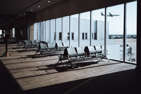 stock image Empty airport terminal seating area with rows of chairs facing large windows, through which a plane can be seen taking off. Sunlight streams in, casting shadows on the floor