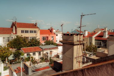 Kırmızı kiremitli çatıları, görünür antenleri ve gür bahçeleri olan geleneksel konut binalarının yer aldığı Lizbon şehir manzarası. Açık mavi bir gökyüzü ve güneş ışığı kentsel mahalleyi aydınlatıyor.