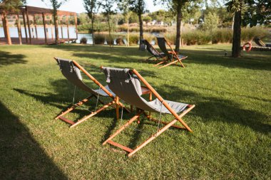 Çimenli bir çimenlikte gri kumaş koltuklu iki ahşap şezlonga ait bir fotoğraf. Arka planda, ağaçlar ve yeşilliklerle çevrili ahşap bir güvertenin yanında daha fazla sandalye görülebilir.