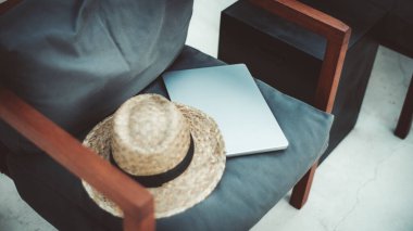 Hasır bir şapka ve ahşap kollukları olan yastıklı bir sandalyeye yerleştirilmiş kapalı bir dizüstü bilgisayar. Ayarlar gündelik ve rahat, iş ve eğlence kavramları için mükemmel.