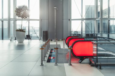 Modern havaalanı içinde kırmızı bir yürüyen merdiven, metal bariyerler ve güneş ışığı altında büyük bir saksı bitkisi var. Temiz, geniş alanda cam duvarlar ve parlak, çağdaş bir tasarım var.