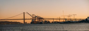 Panoramic low-key shot of Lisbon's harbor at sunset, featuring the 25 de Abril Bridge and various cranes in the port. The warm hues of the setting sun enhance the industrial and architectural elements clipart