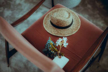 Hasır bir şapka, küçük bir buket kurumuş çiçek ve kırmızı döşemeli bir sandalyeye yerleştirilmiş beyaz bir not kartı. Sahne sığ bir alan derinliğiyle rahat ve aydınlık.