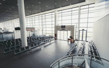 Geniş, modern bir havaalanı, sayısız siyah sandalye ve geniş cam pencereli doğal ışığa izin veren bir bekleme alanı. Alan bir bilgi masası ve dijital ekran tahtasını içerir; geniş açı