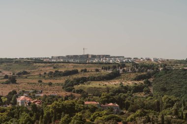 Ufukta dağınık evler ve inşaat halinde olan tepelik bir manzaranın panoramik görüntüsü. Yeşil ağaçlar ve bitki örtüsü açık bir gökyüzünün altında