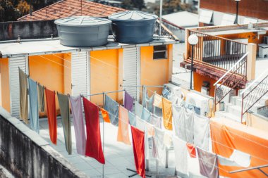 Elbiseler ve havlular bir binanın çatı katındaki çamaşır sahasında kurumaya bırakılmış. Boşlukta parlak turuncu ve sarı duvarlar, beyaz panjurlar, çatıda su tankları ve açık hava merdivenleri yer alıyor.