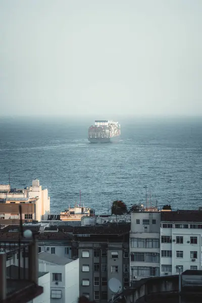 stock image A vertical telephoto of a large container ship sails into the ocean, viewed from a cityscape with residential buildings in the foreground. The ship's journey against a calm sea and clear sky