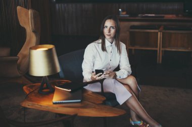 Beyaz gömlekli ve açık mor etekli profesyonel bir kadın modern ofis ortamında elinde akıllı bir telefonla oturuyor. Bir dizüstü bilgisayar, defter, lamba onun yanındaki ahşap masada; sığ alan derinliği